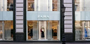 Fachada de loja da Zara com vitrine envidraçada, manequim exibindo coleção de roupas no centro e interior iluminado, representando marca de moda global e varejo de vestuário.